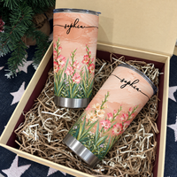Two personalized birth flower tumblers displayed inside an open gift box filled with shredded paper filler.