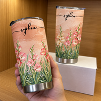 A hand holding a personalized birth flower tumbler in 20oz size with peach background and name Sophia next to another