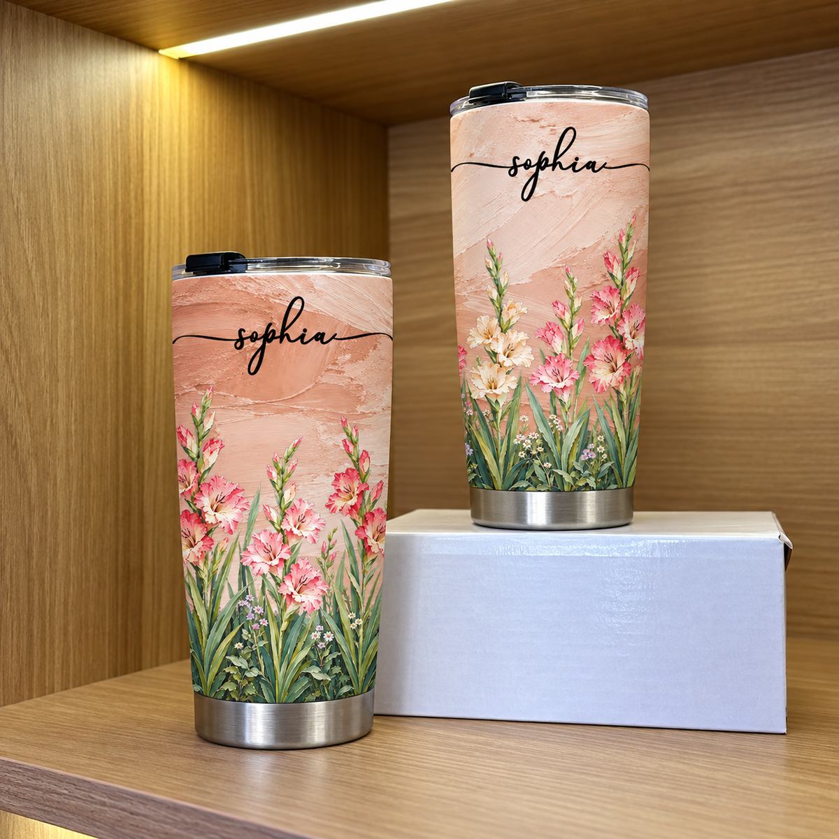 A 20oz and a 40oz personalized birth flower tumbler displayed together on a white box inside a wooden shelf.
