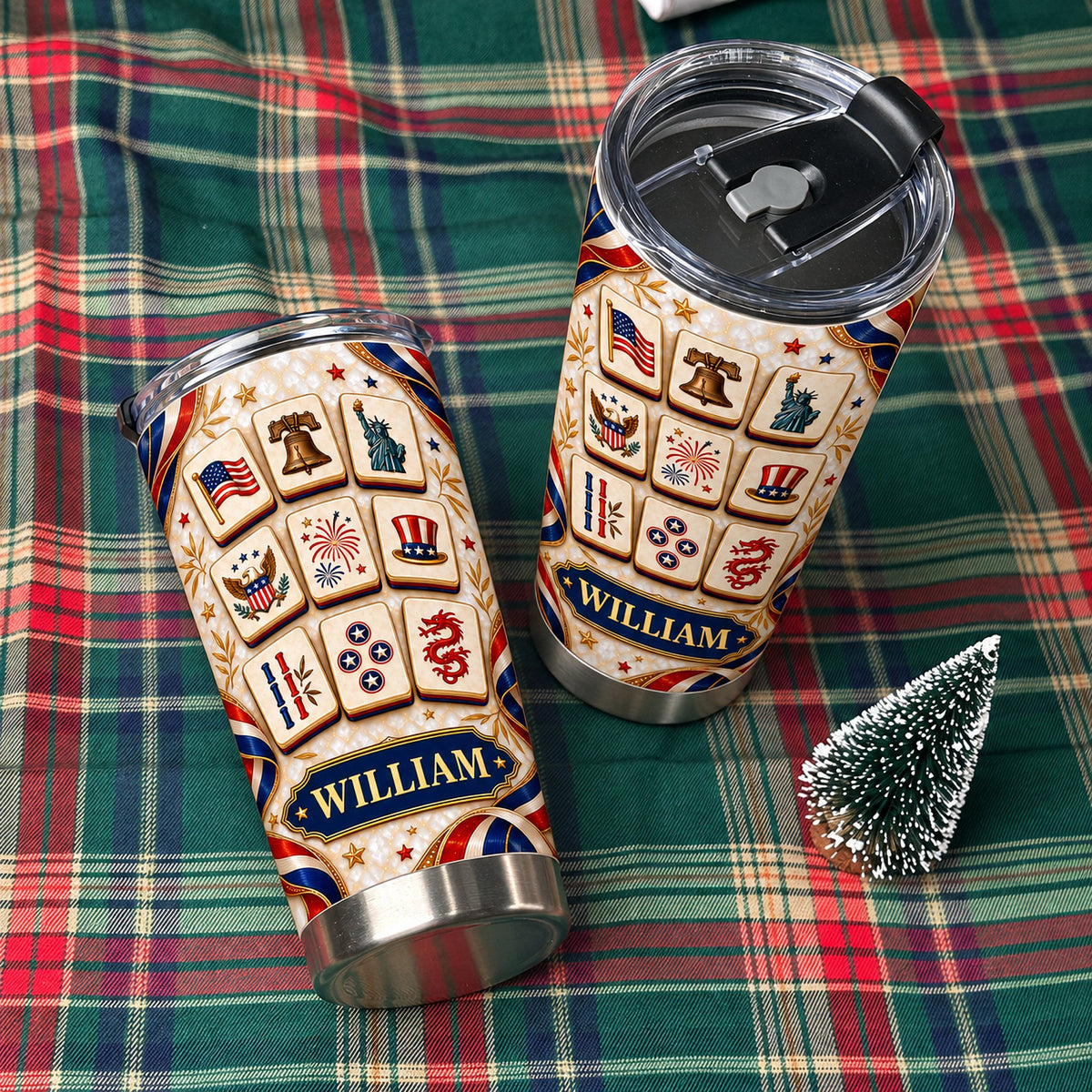 Two personalized patriotic mahjong tiles tumblers with black lids resting on a green plaid blanket next to a tiny pine tree.