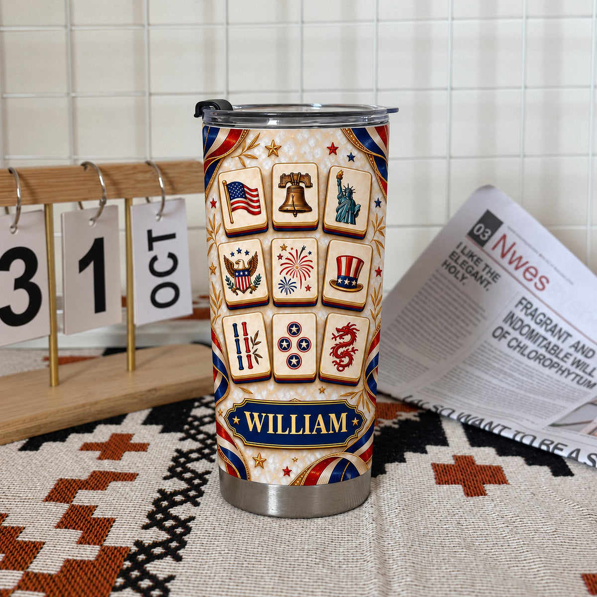 Personalized patriotic mahjong tiles tumbler standing on a patterned rug next to an October calendar and newspaper.