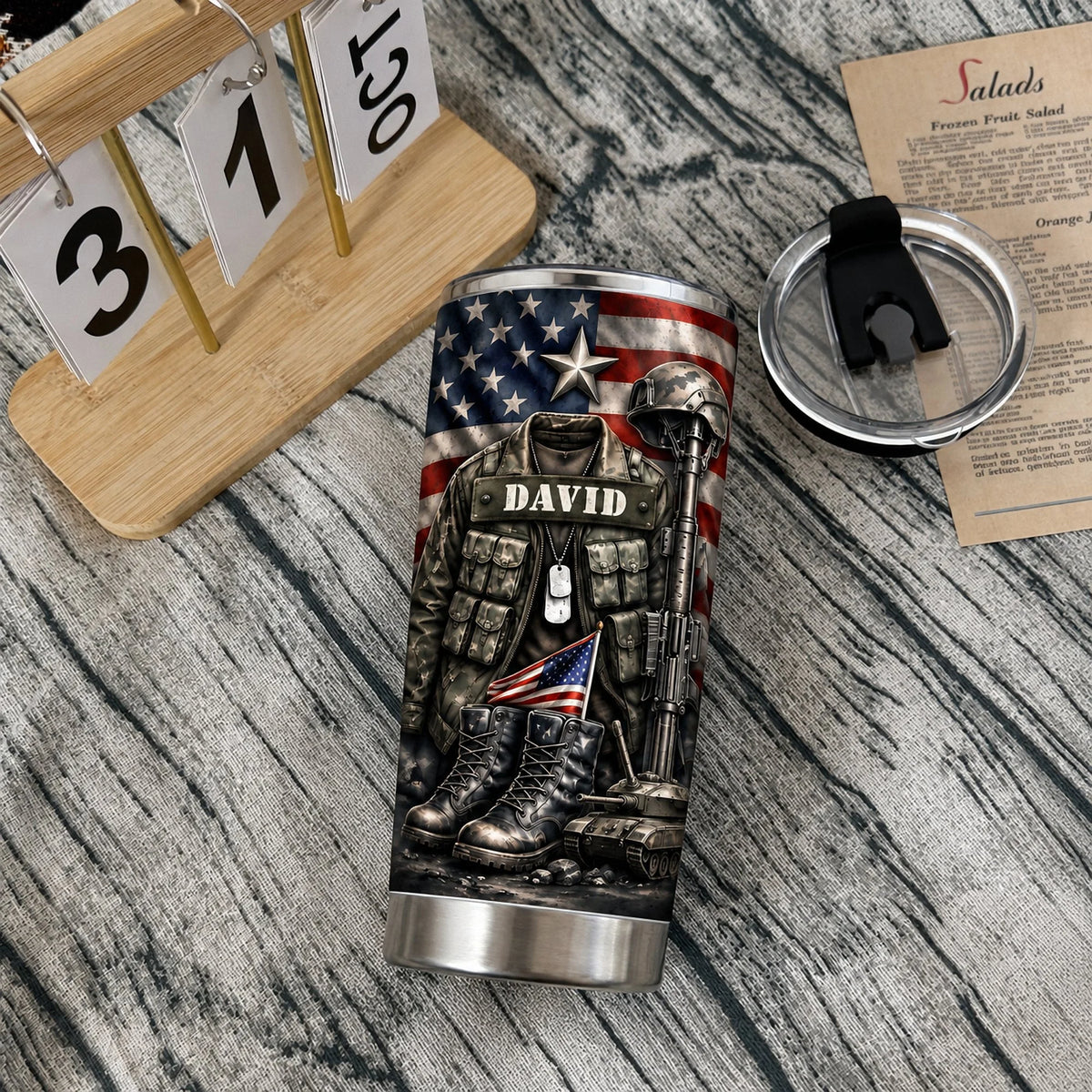 A stainless steel army veteran gift tumbler with a lid next to a calendar stand and a note on a wooden table.