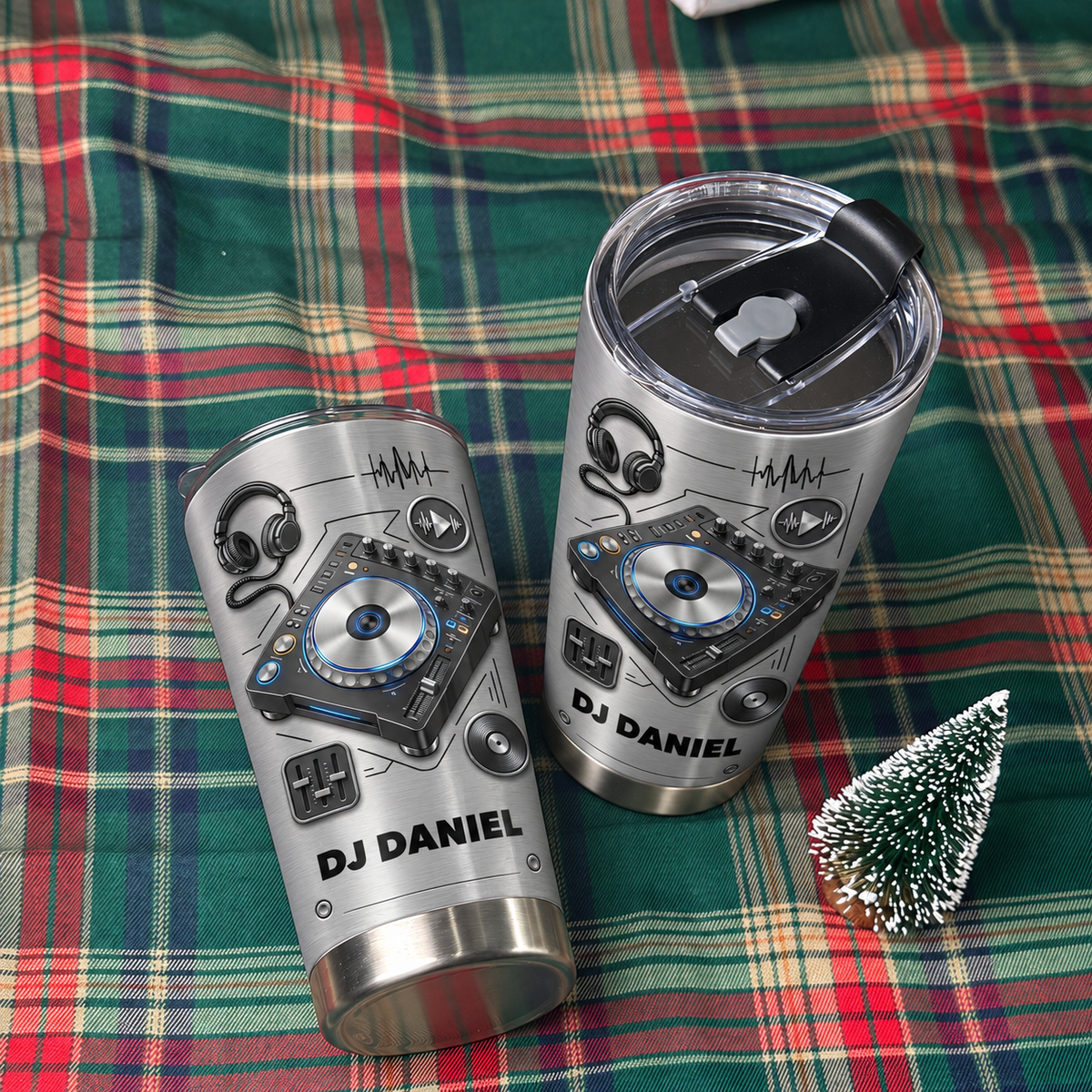 Two personalized DJ tumblers resting on a green and red plaid blanket beside a miniature tree.