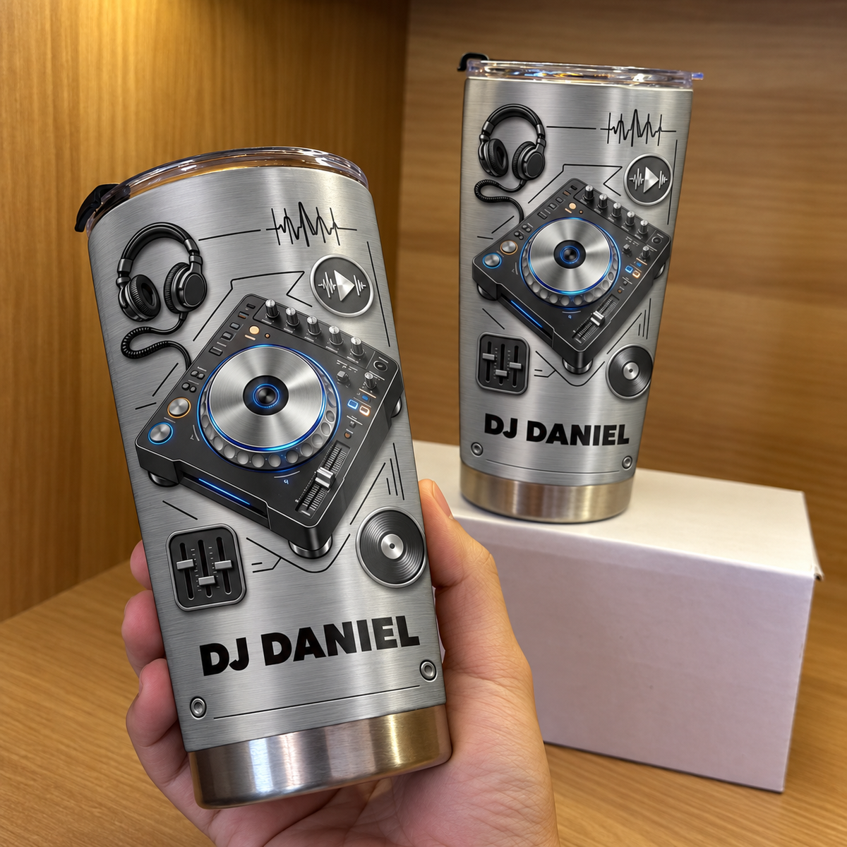A person holding a personalized stainless steel DJ tumbler with a second tumbler on a white box behind.