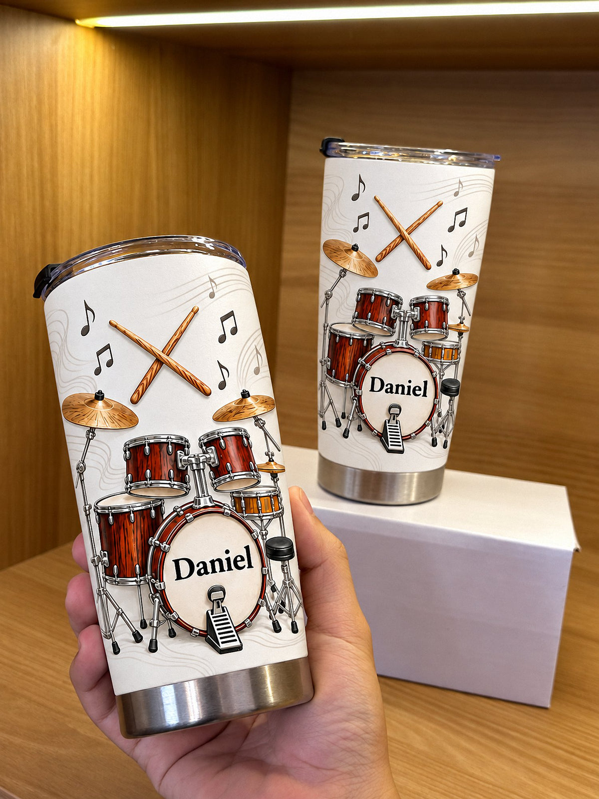 Two custom name tumblers featuring drum kit graphic and Daniel text, displayed upright on wooden shelf with lighting.