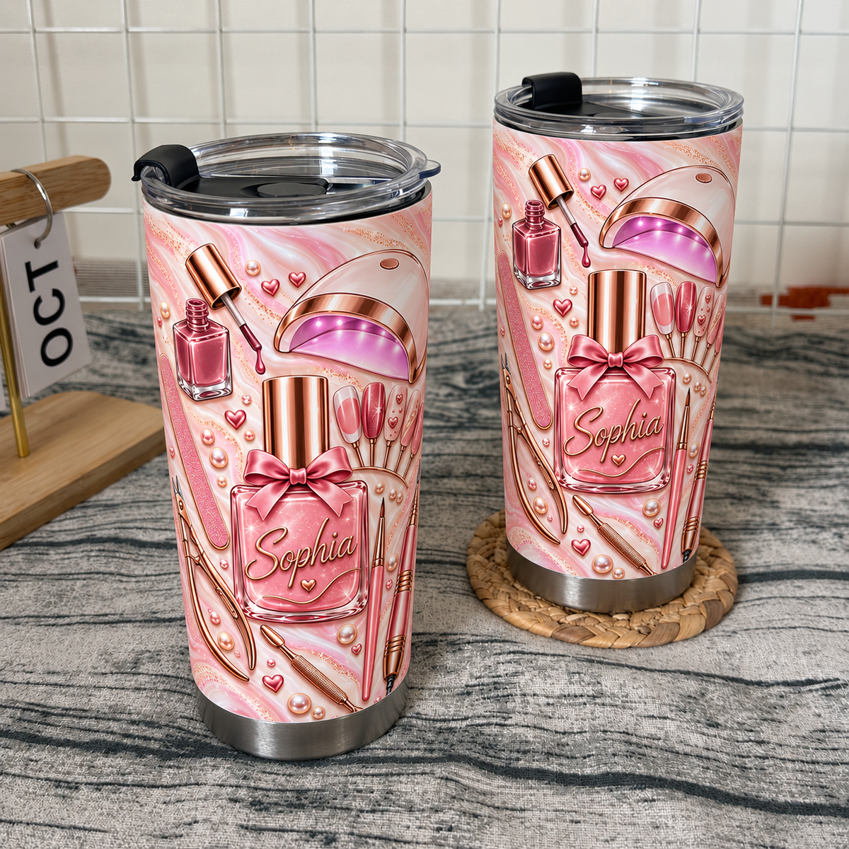 Two stainless steel drinkware tumblers featuring pink marble and manicure tool illustrations stand side by side on a woven