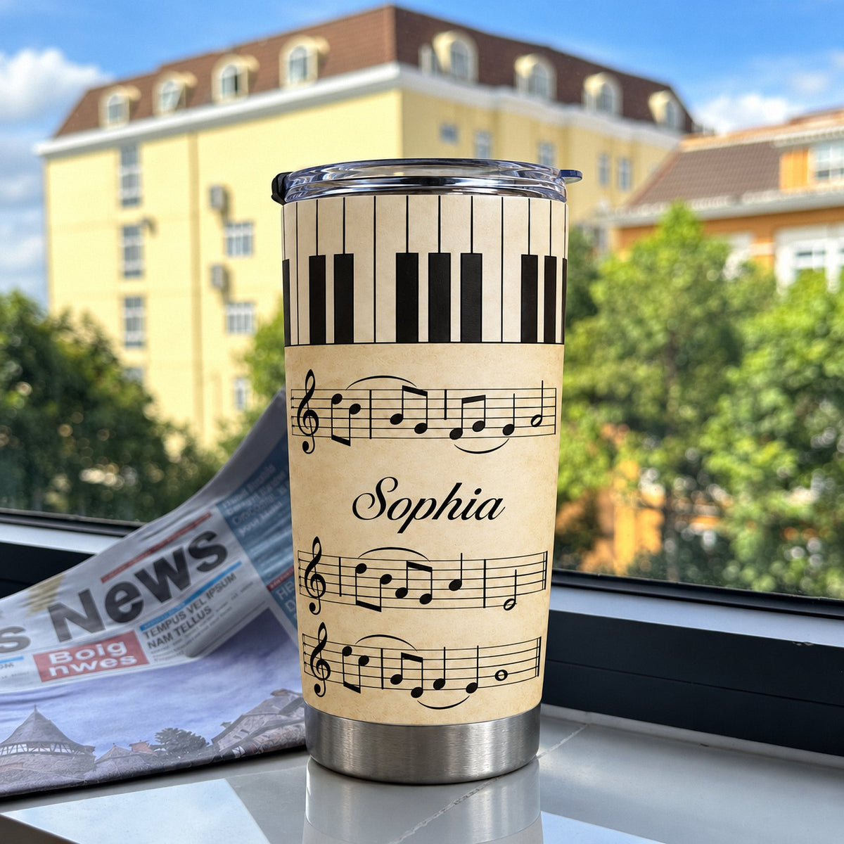 A stainless steel tumbler with vintage piano sheet music design and the custom name Sophia, standing on a windowsill with a