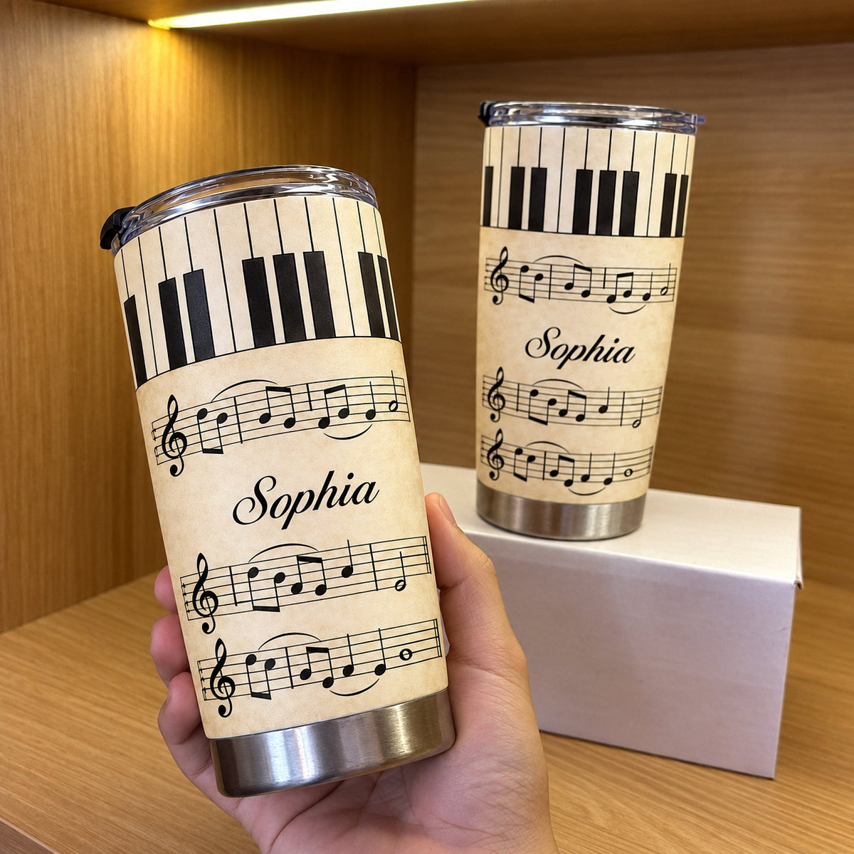 Hand holding a personalized piano sheet music tumbler next to a second tumbler on a white box against a warm wooden shelf