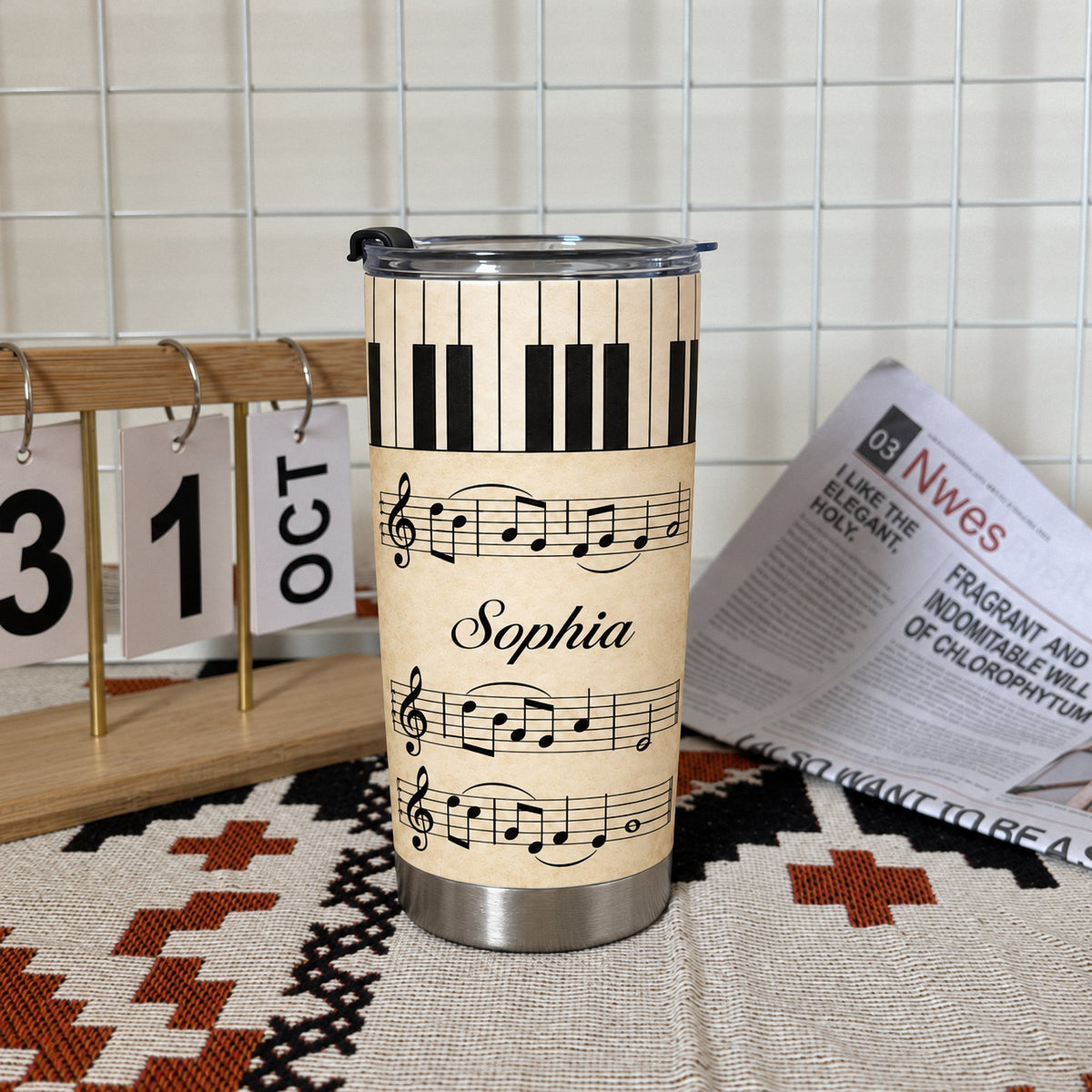 Personalized piano sheet music tumbler standing on a patterned mat next to a small wooden calendar and a newspaper on a white