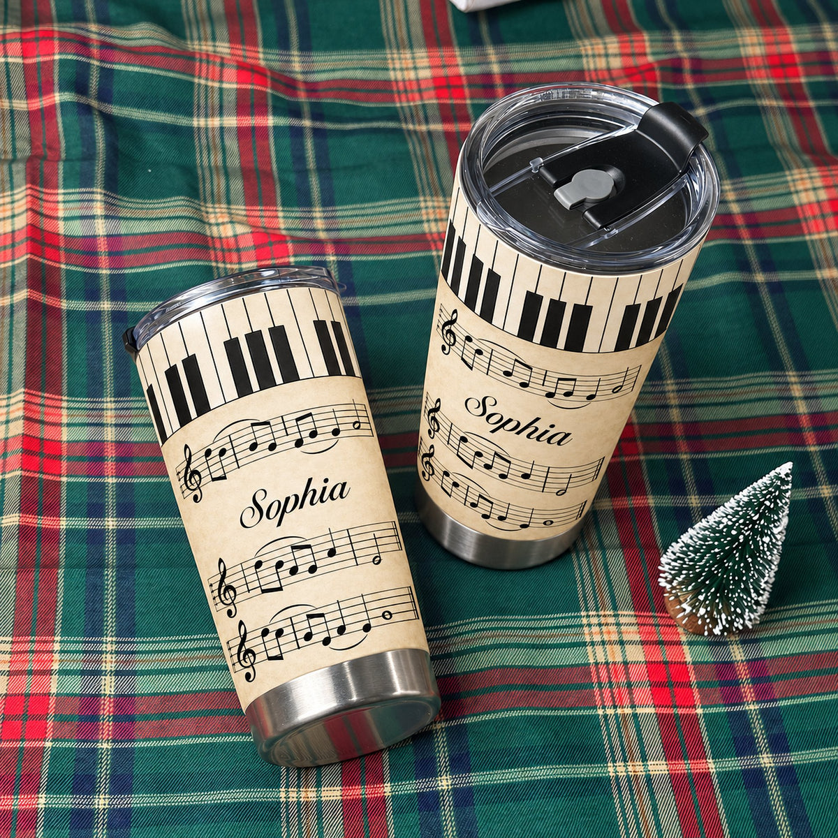 Two stainless steel music tumblers with vintage piano keys and sheet music design resting on a red and green plaid fabric