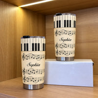 Personalized piano sheet music tumbler standing upright beside a second tumbler on a white box inside a wooden display shelf.