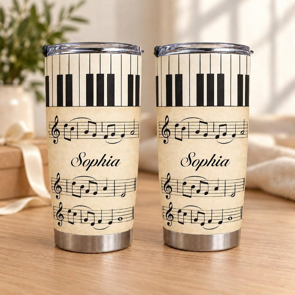 Two personalized piano sheet music tumblers standing on a wooden surface with a soft-focus plant and white ribbon background.