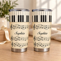 Two personalized piano sheet music tumblers standing on a wooden surface with a soft-focus plant and white ribbon background.