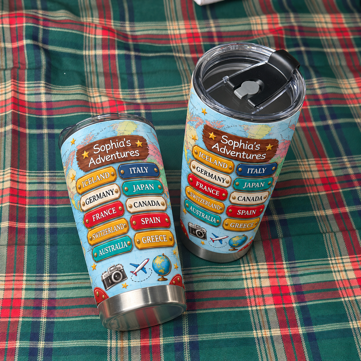 Two personalized travel map tumblers resting sideways on a red and green plaid fabric surface.
