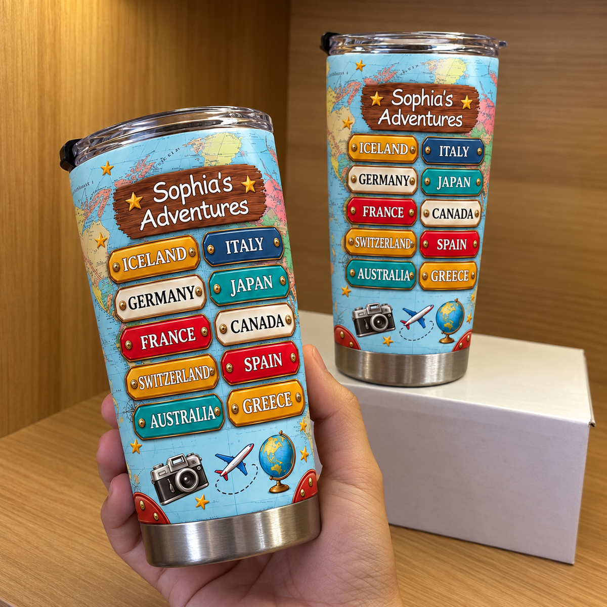 Hand holding a personalized travel map tumbler with luggage tag labels next to another on a white box.