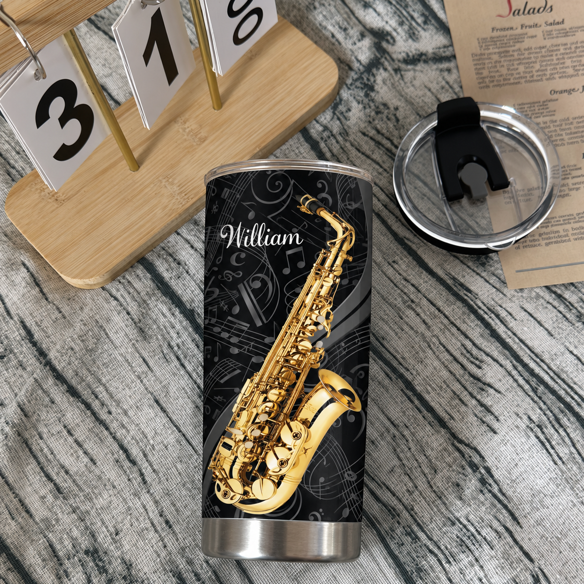 Saxophone musician tumbler featuring jazz art, set next to a clear lid and a wooden calendar on a rustic table.