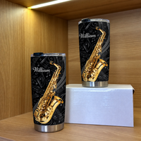 Two black and gold custom name tumblers with saxophone designs displayed on a white box against a wooden backdrop.