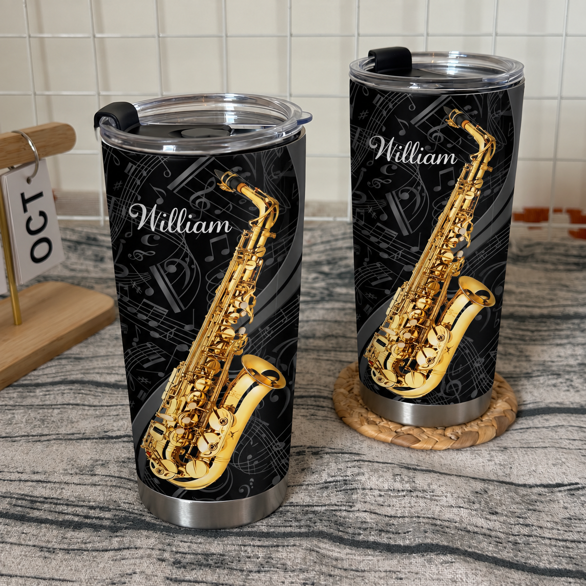 Two custom saxophone musician tumblers with black and gold prints displayed on a textured grey surface with a calendar.