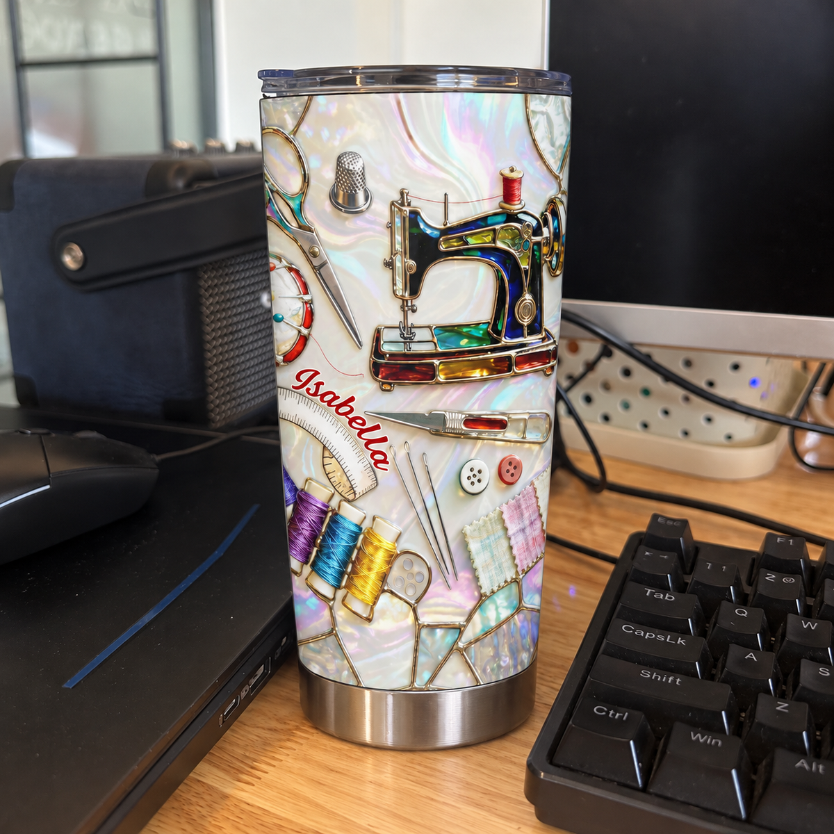 A custom name tumbler with sewing machine design stands upright on a wooden desk next to a computer keyboard.