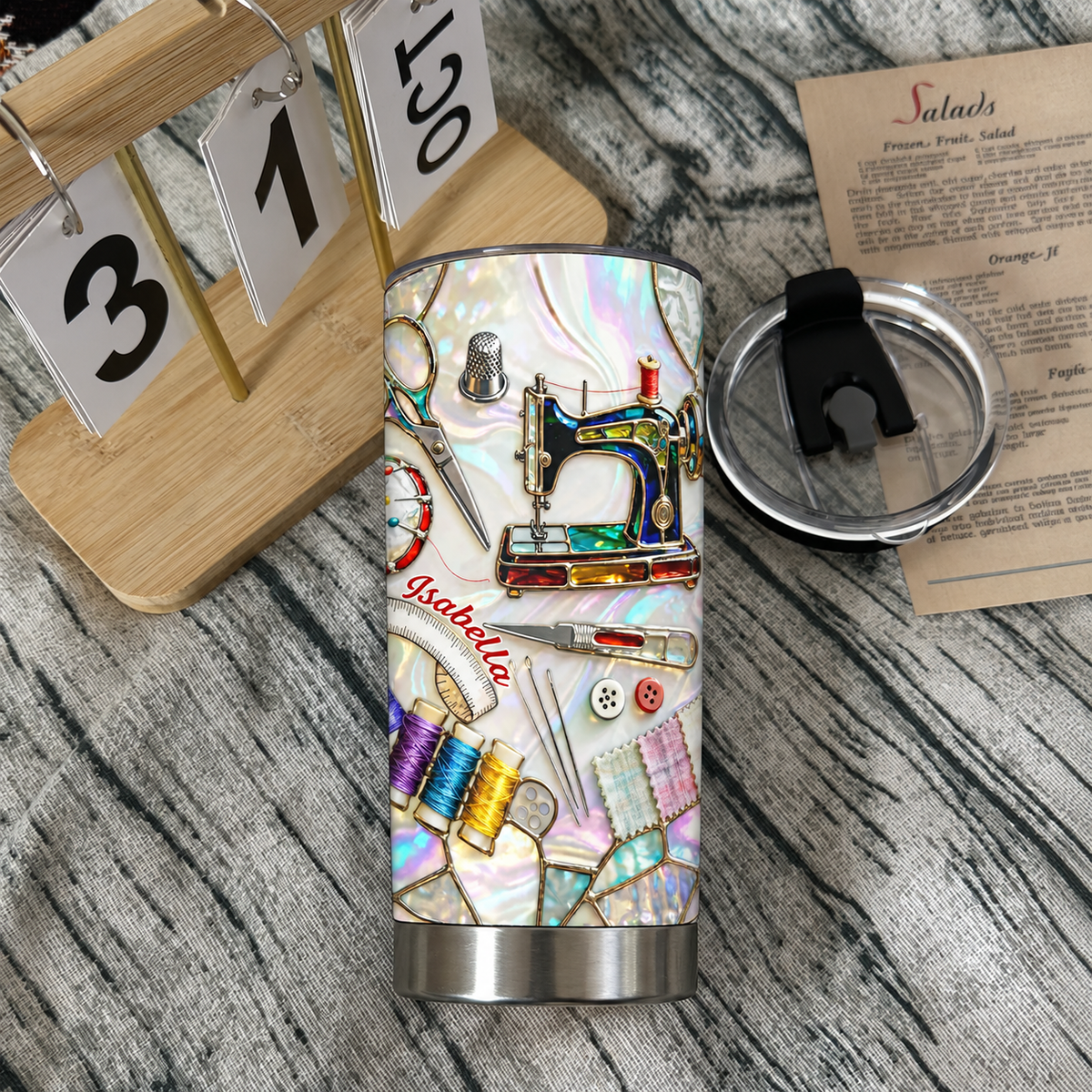 Stainless steel insulated tumbler with sewing machine design resting on a wooden surface next to a wooden desk calendar.