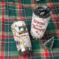 Two custom name tumblers with sewing machine design lying on a green plaid blanket next to a small holiday tree.
