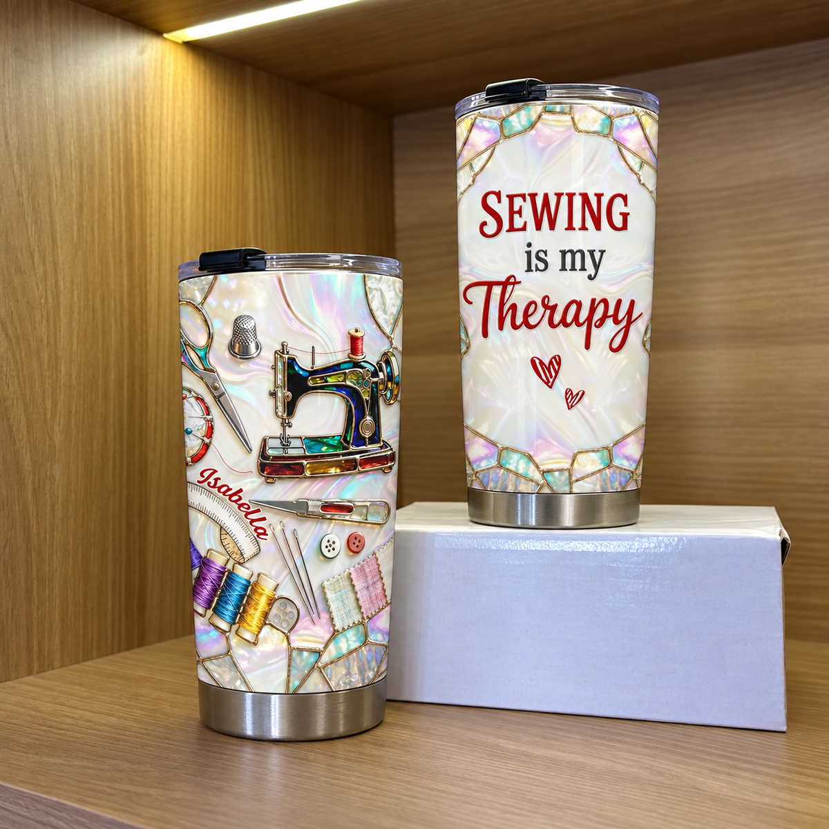 Two insulated stainless steel tumblers with sewing machine design displayed on a white box inside a wooden cabinet.
