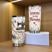 Two insulated stainless steel tumblers with sewing machine design displayed on a white box inside a wooden cabinet.