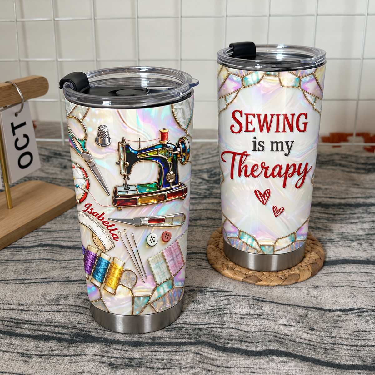 Insulated stainless steel tumbler with custom name and sewing machine design standing beside a matching tumbler on a rustic