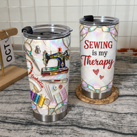 Insulated stainless steel tumbler with custom name and sewing machine design standing beside a matching tumbler on a rustic