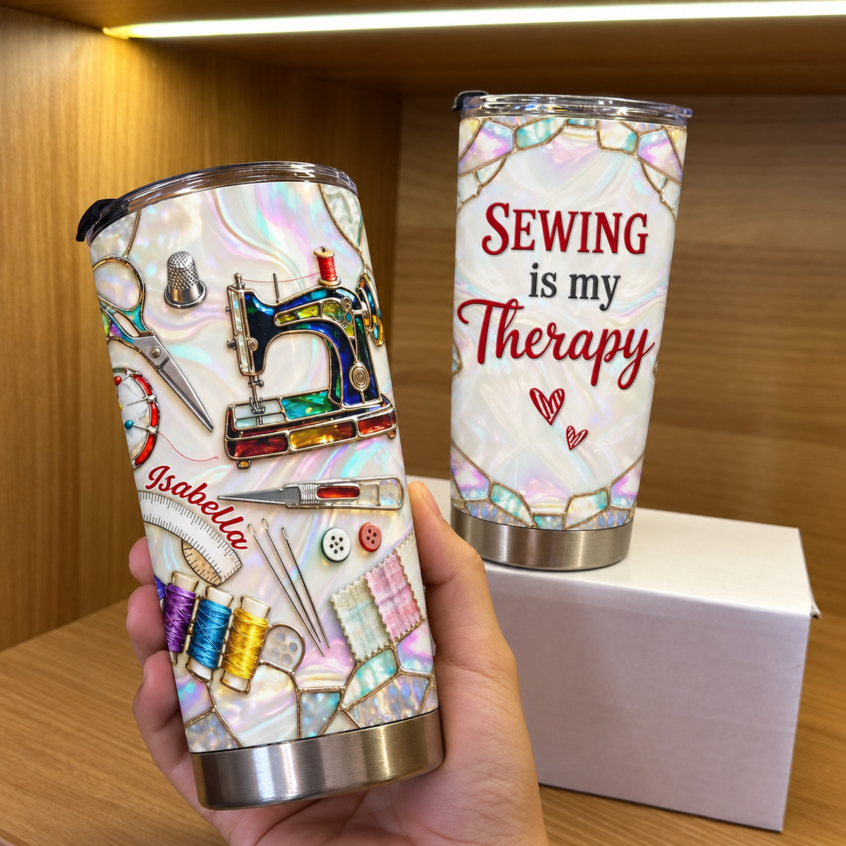 Hand holding a custom name tumbler featuring a vintage sewing machine design, with a second tumbler on a box behind it.