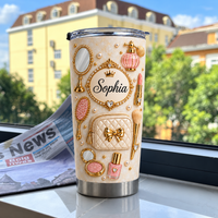 Personalized gold pearl vanity tumbler displayed on a windowsill with a newspaper and blurred building background.