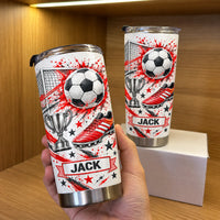 A hand holds a red soccer tumbler personalized custom name next to a second cup on a white box in front of wood shelves.