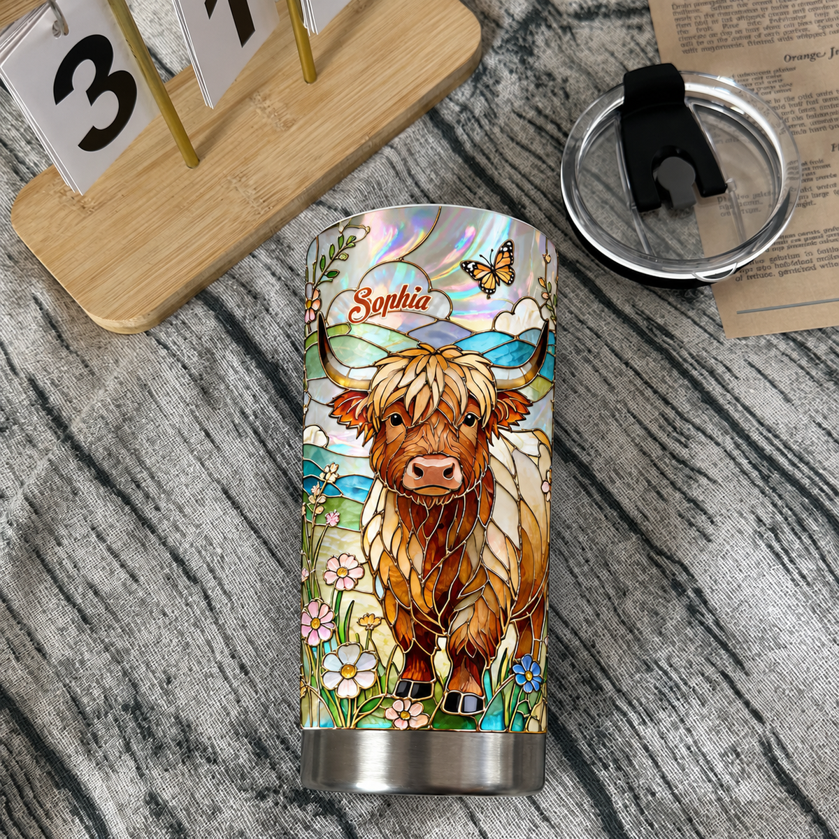 Stainless steel tumbler cup with stained glass Highland cow design and custom name banner beside lid and calendar on wooden