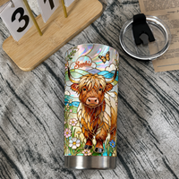 Stainless steel tumbler cup with stained glass Highland cow design and custom name banner beside lid and calendar on wooden