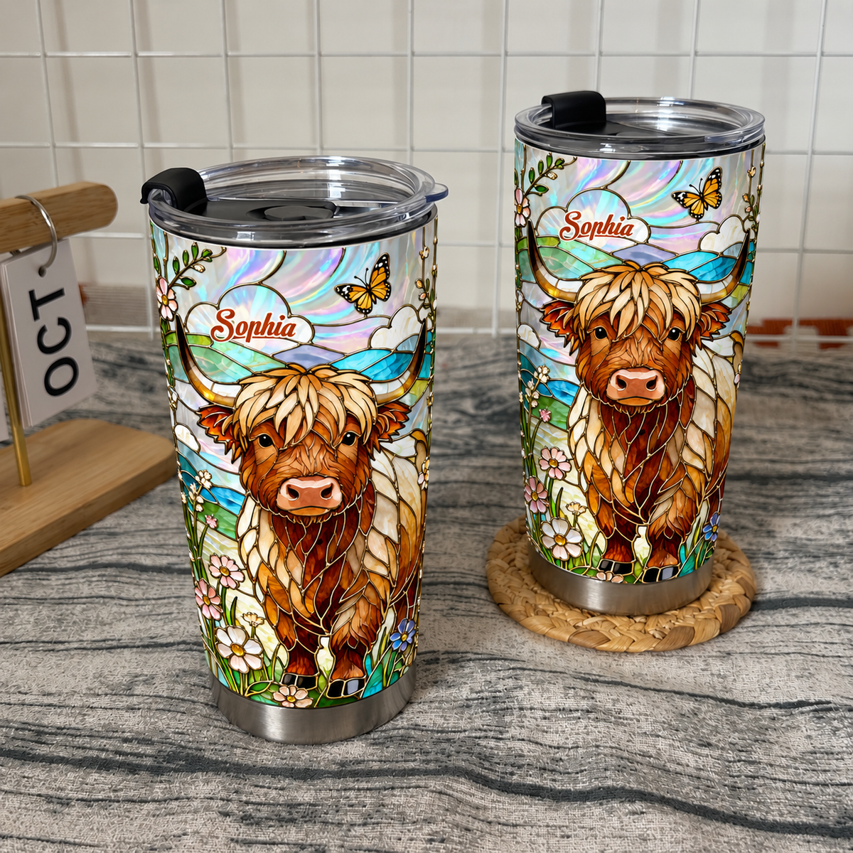 Two custom name tumbler cups with Highland cow stained glass art on wooden surface with calendar and woven coaster.