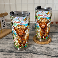 Two custom name tumbler cups with Highland cow stained glass art on wooden surface with calendar and woven coaster.