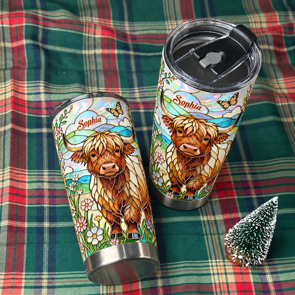 Two stainless steel tumbler cups with Highland cow and floral art displayed on green plaid blanket with mini Christmas tree.