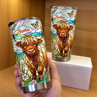 Hand holding a custom name tumbler featuring stained-glass Highland cow design, with second cup on white box in wooden