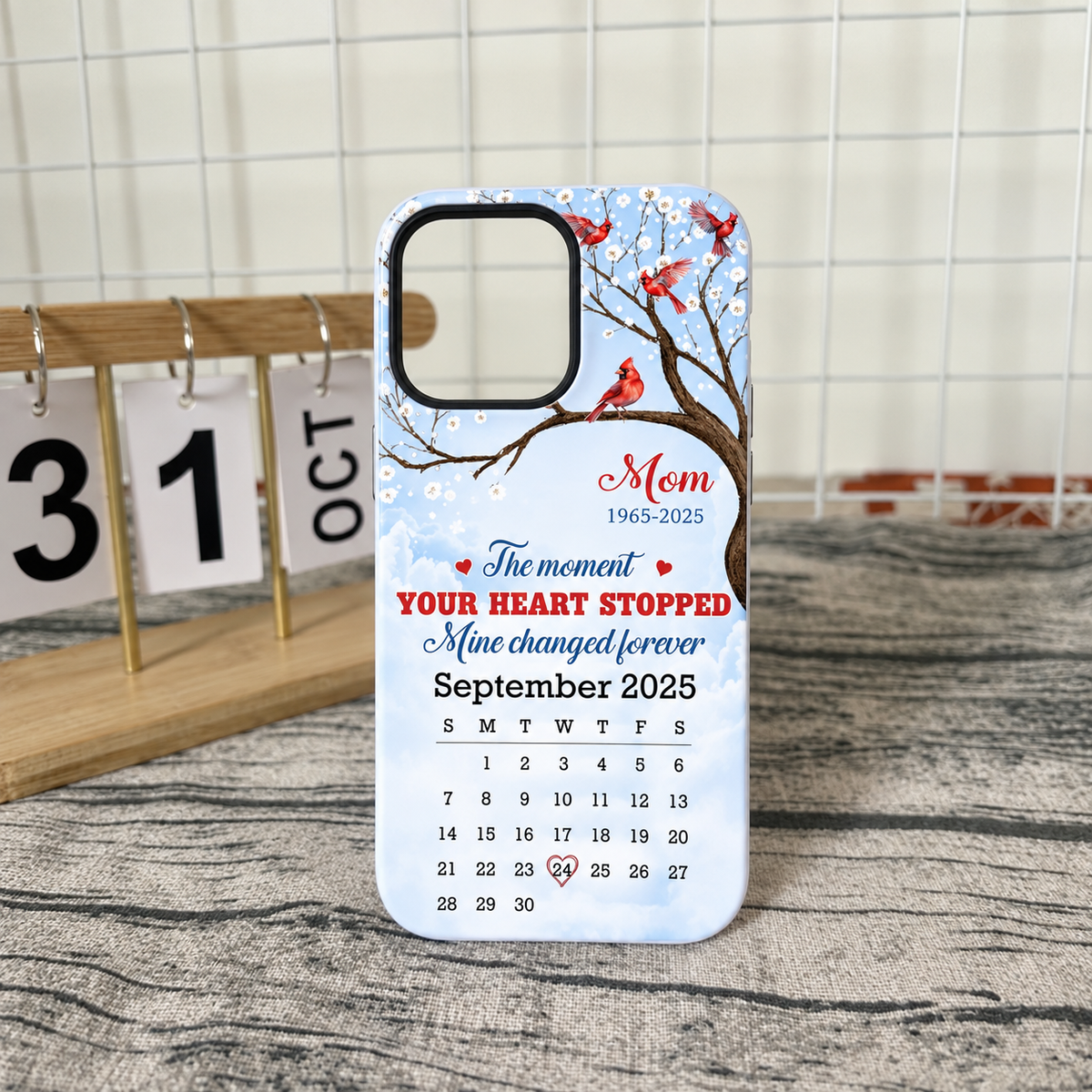 Memorial Cardinal Phone Case standing upright on a grey surface in front of a white wire grid and wooden calendar block.