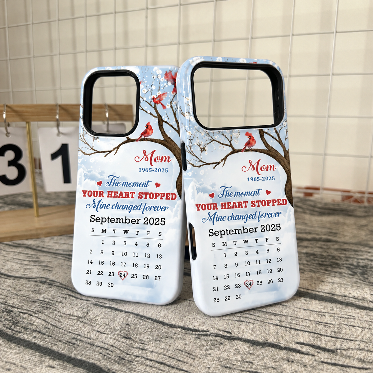 Two Memorial Cardinal Phone Cases displayed upright on a grey surface in front of a white wire grid backdrop.