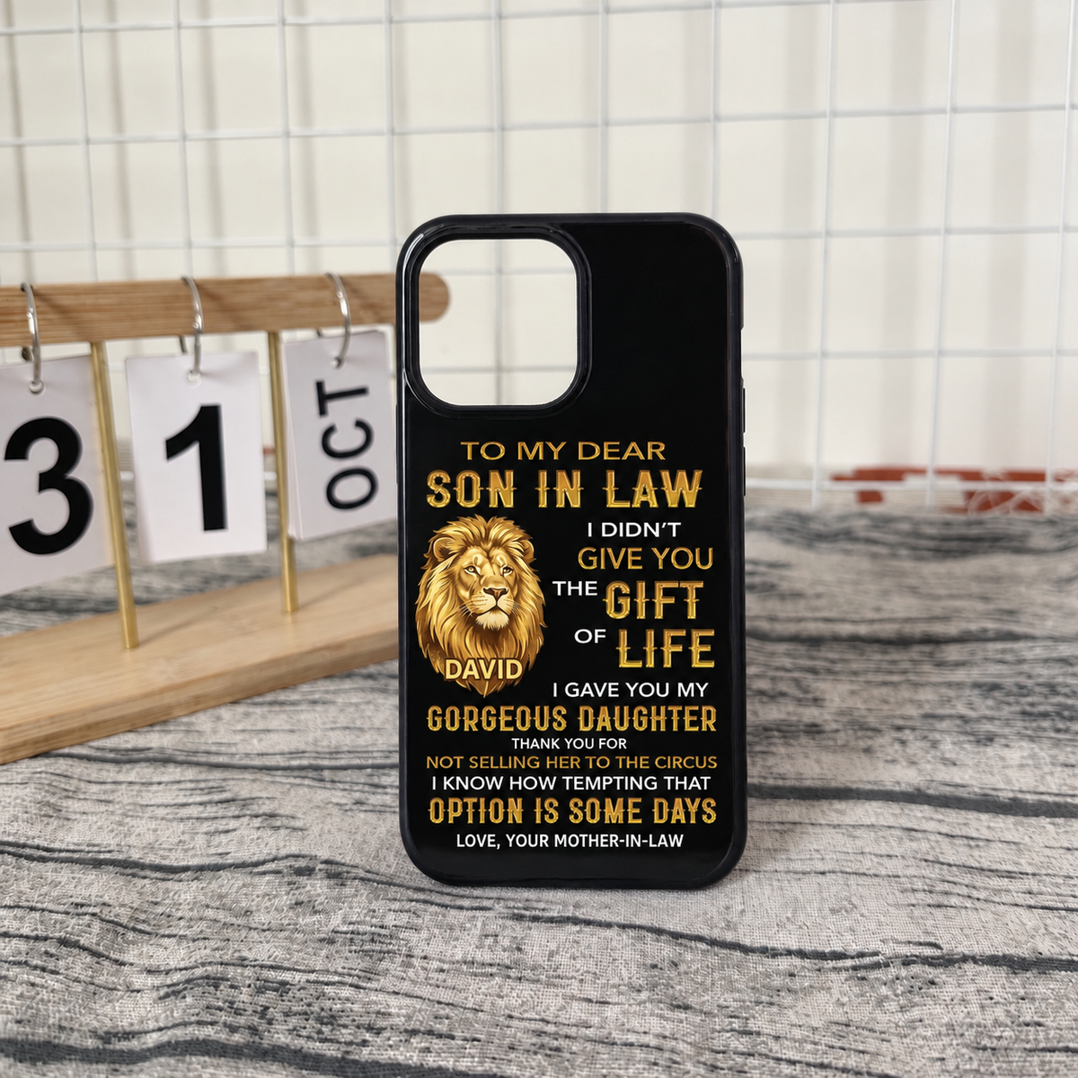 A black iPhone tough case with a gold lion and witty family message standing upright on a textured table near a calendar desk