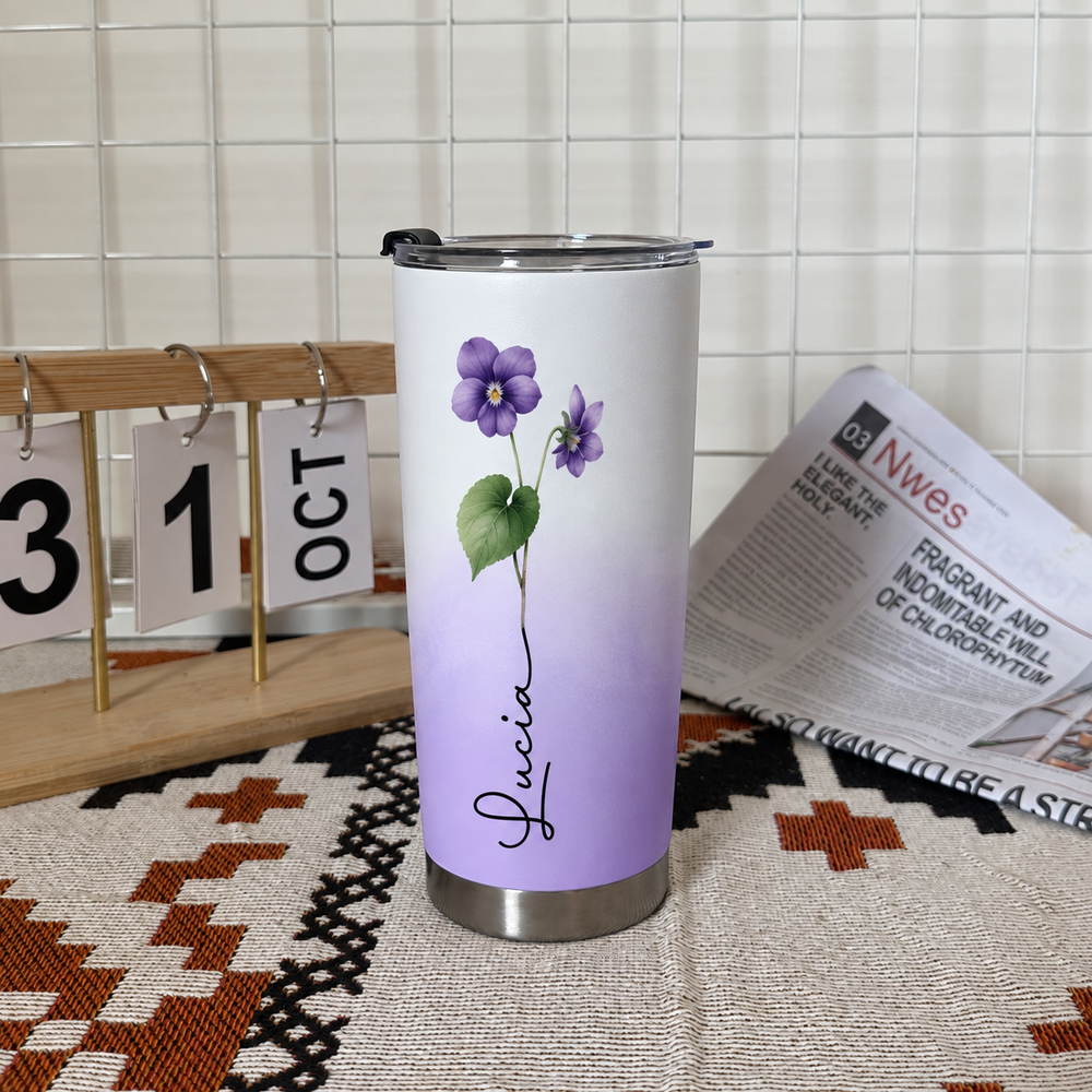 Custom pansy flower tumbler with recipient's name standing on a patterned mat near a calendar and newspaper.