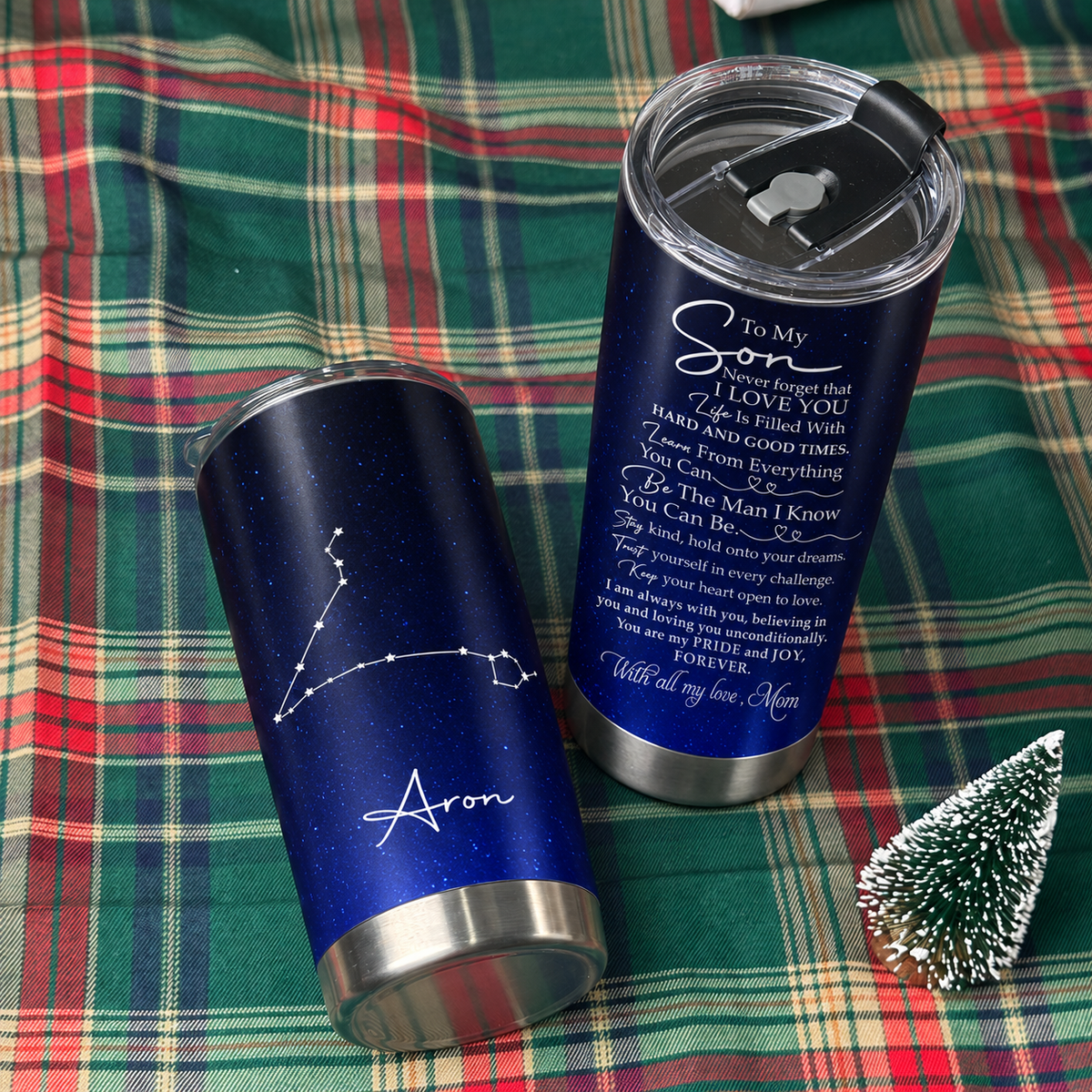 Two stainless steel tumbler cups with inspirational text rest on a red plaid blanket beside a tiny tree.