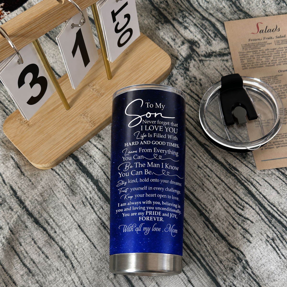 A stainless steel tumbler cup featuring an inspirational message lies flat beside its clear lid and a wooden calendar.