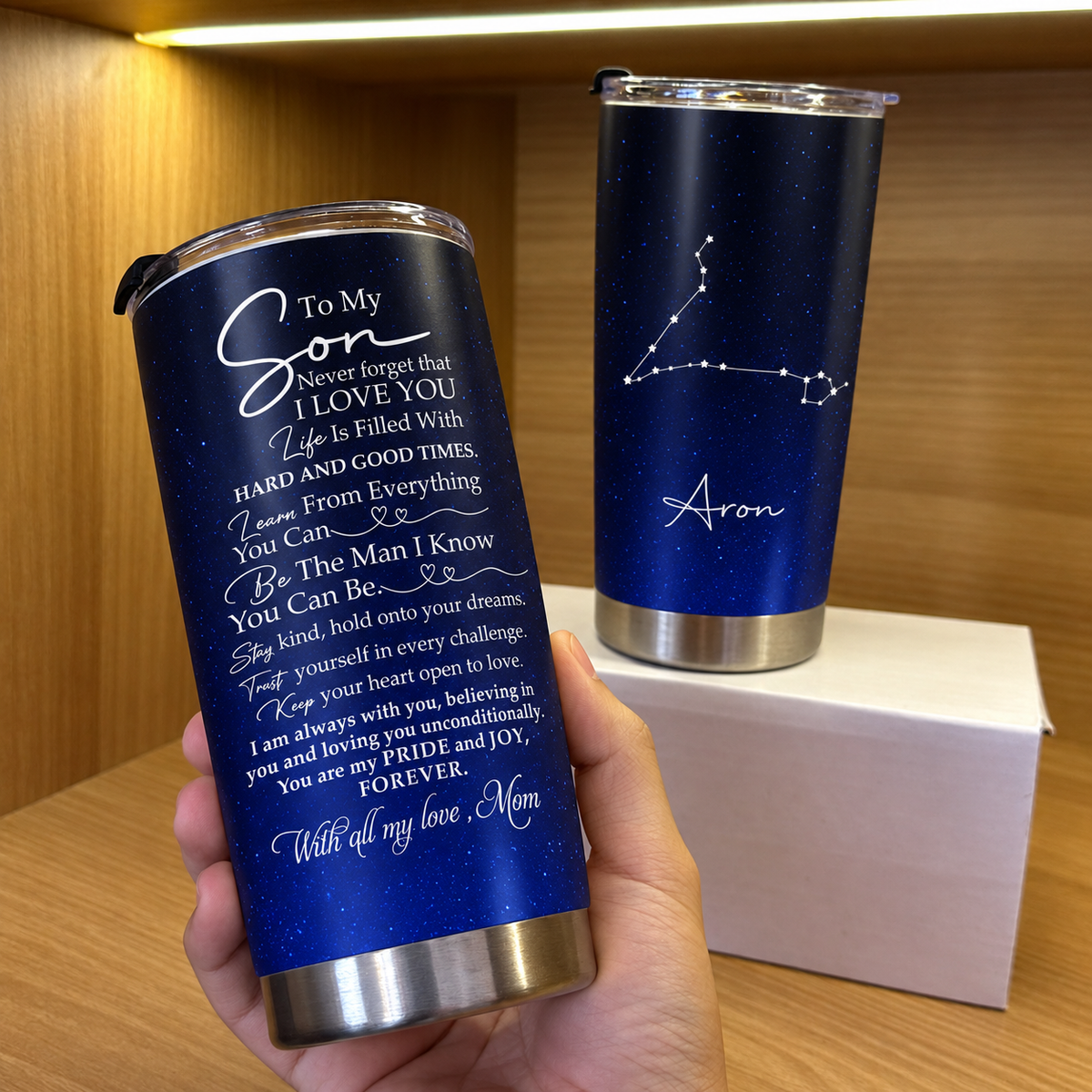 A hand holds a stainless steel tumbler with a graduation gift quote while another cup stands on a box behind it.