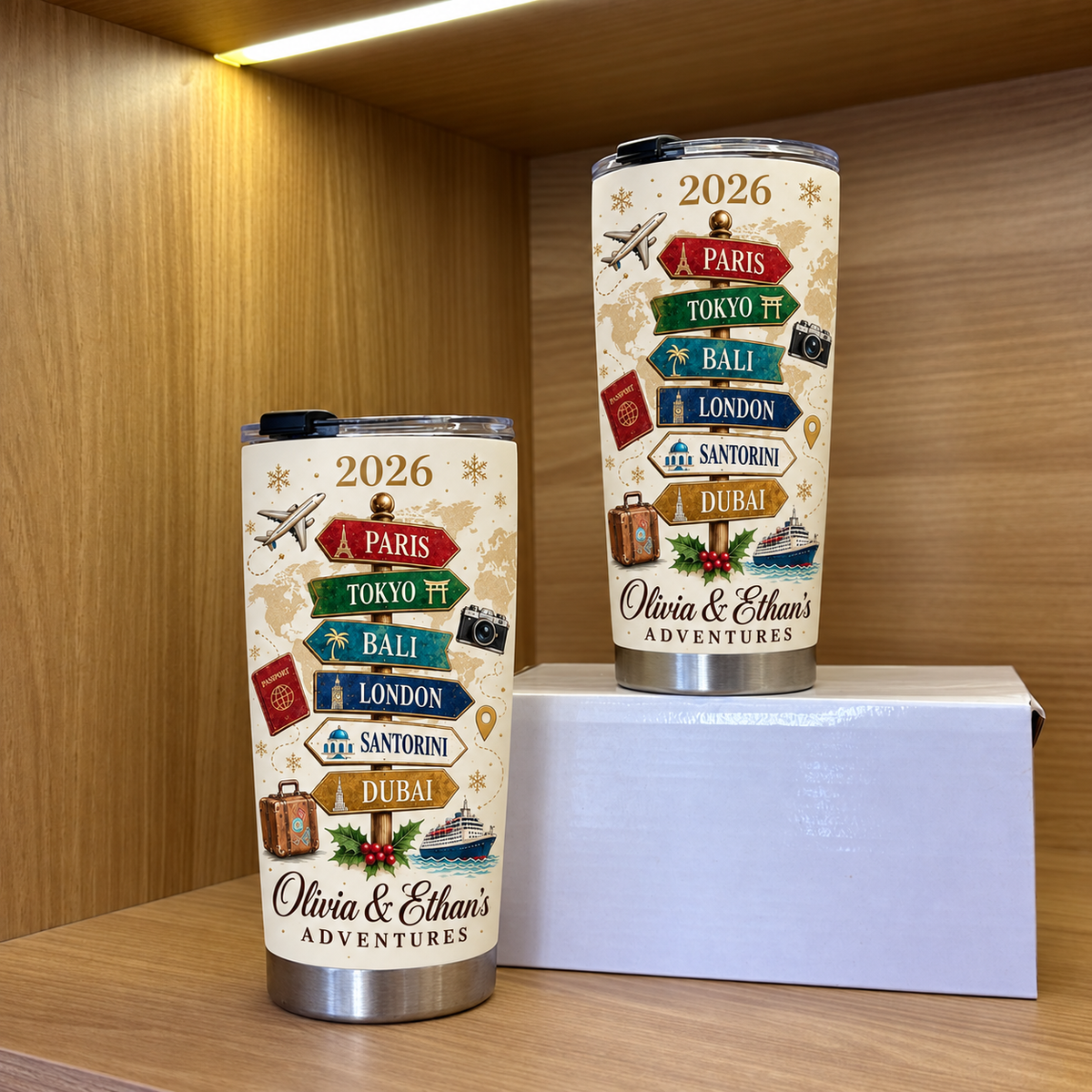 Two stainless steel travel cups featuring a vintage travel signpost design displayed on a wooden shelf next to a white box.