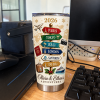 A custom stainless steel travel tumbler featuring a vintage travel signpost and couple names on a wooden desk near a