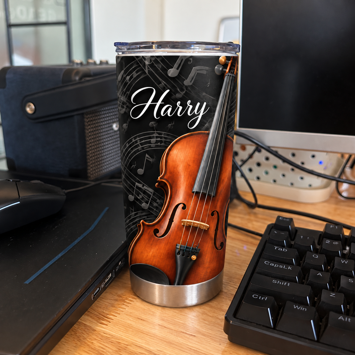 Custom violin music tumbler placed on a wooden desk next to a computer keyboard and monitor in an office.