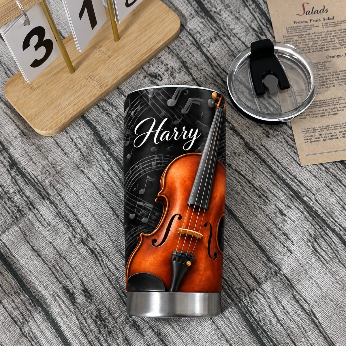 Personalized violin insulated travel mug with sheet music design and clear lid beside a calendar on a wooden table.