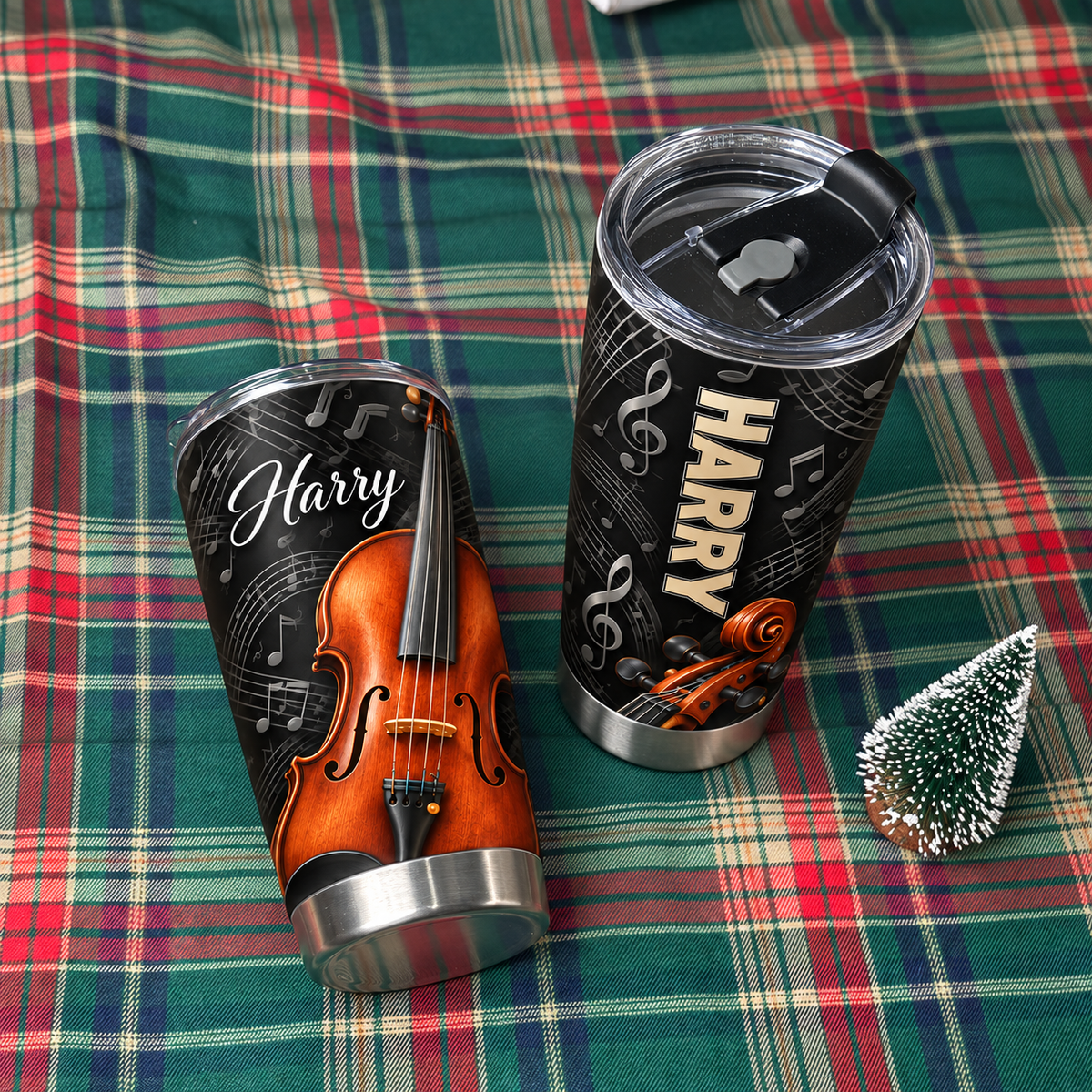 Two personalized violin music tumblers resting on a green and red plaid blanket next to a small decorative pine tree.